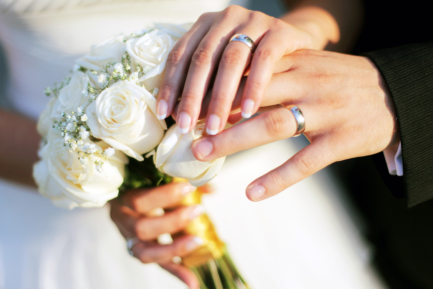 A newlywed couple showing off their rings and holding a bouquet of white roses.