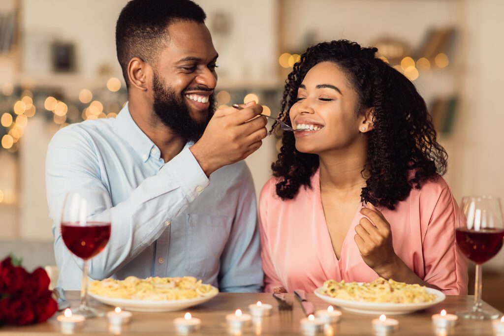 A smiling man feeding his date pasta at a romantic restaurant.